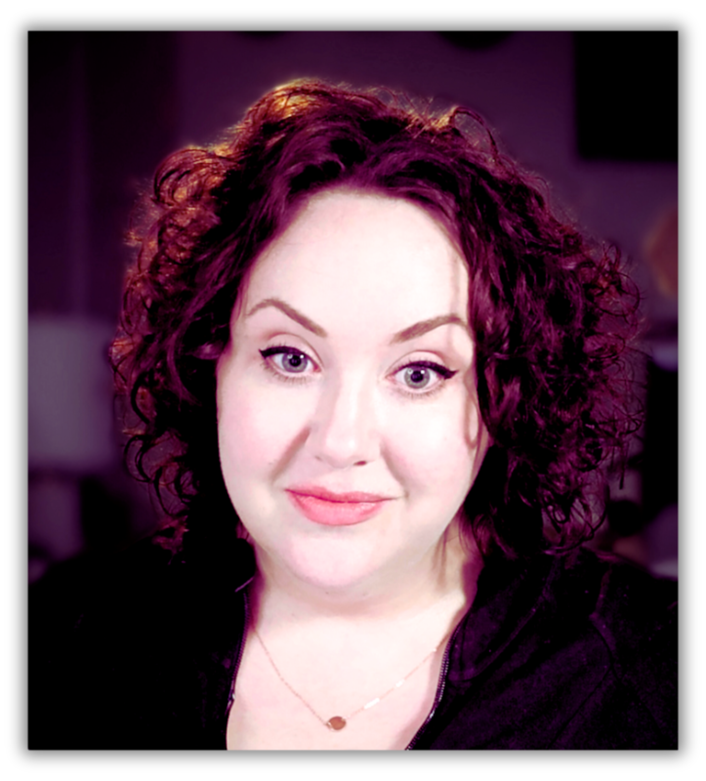 alt="White woman with dark brown curly hair, and green eyes wearing winged eyeliner, a light berry lipstick, gold necklace, and black cardigan.'